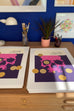 Two printed art sheets displayed on a wooden desk, featuring a pixelated pink skull design on a dark purple background with gold circular accents. The sheets are set against a blue wall backdrop with a white desk lamp, paintbrushes in a glass jar, a small potted plant, and framed artwork.